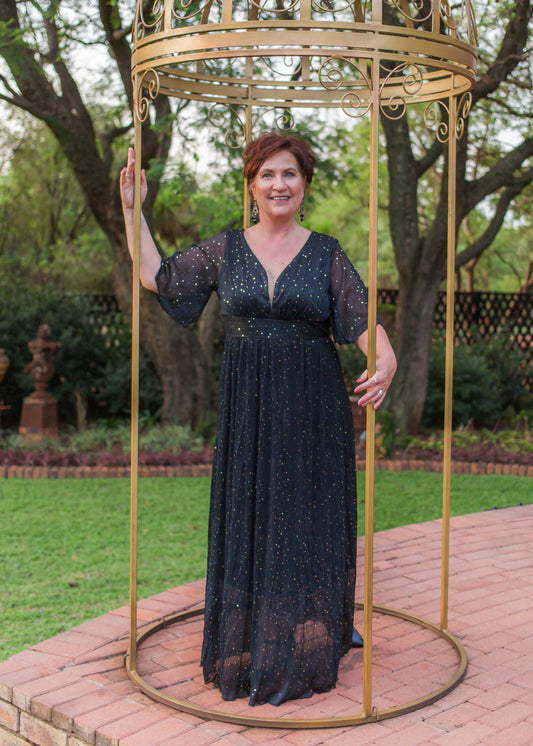 Woman in a black dress standing inside a decorative gold cage in an outdoor setting with trees and a garden.