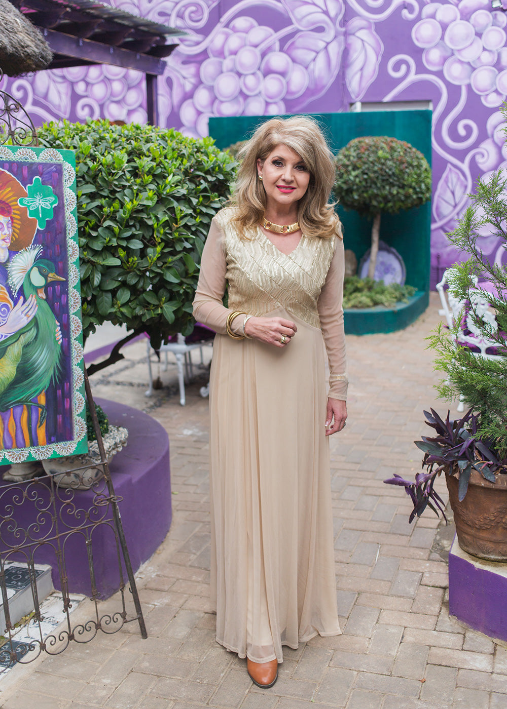Woman in a beige dress standing in front of a colorful mural and plants.