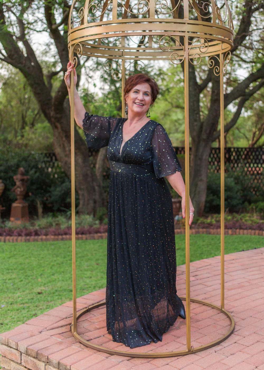 Woman in a black dress standing inside a gold birdcage in an outdoor setting